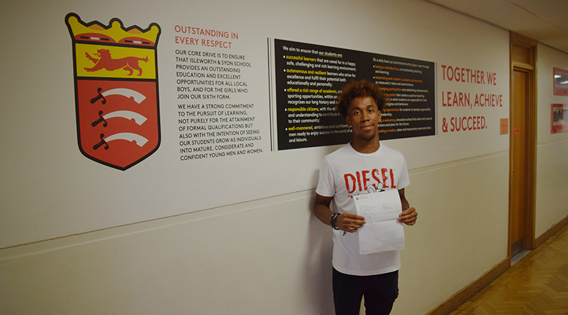 Student Diallo Williams stands holding a piece of paper holding his results. He is standing in a corridor in front of the Isleworth & Syon crest and mission statement.