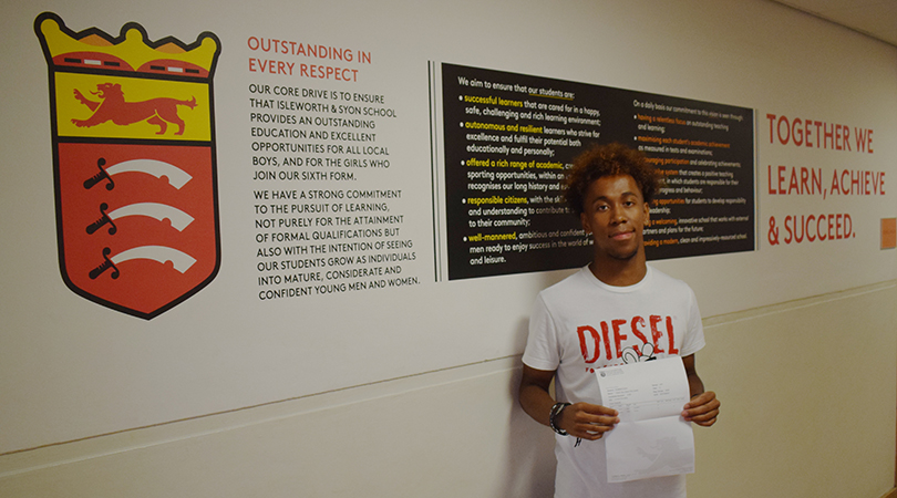 A student holds their results in front of a wall displaying ethos information about Isleworth & Syon School.