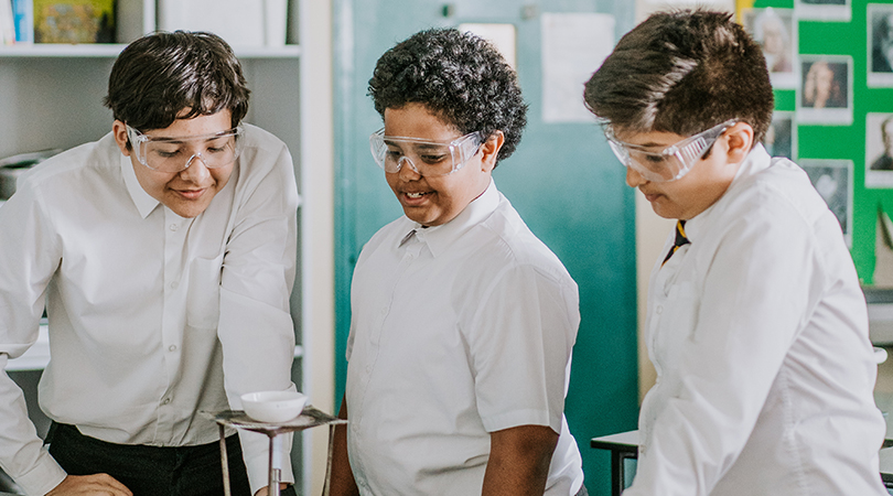 Three students look at a white dish on top of a mesh plate, above a bunsen burner.
