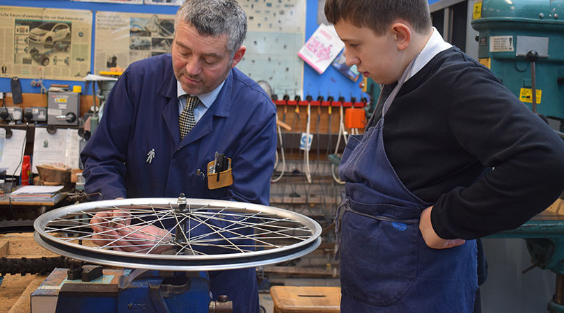 A teacher fixes a bicycle wheel (left) while a student looks on (right).