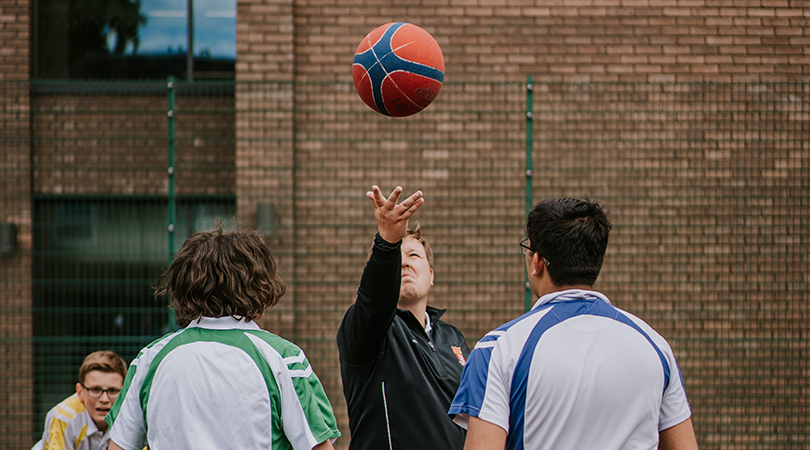 A male teacher throws a basketball in the air. In front of him (back to camera) are two students in house polo shirts waiting to jump for the ball. 