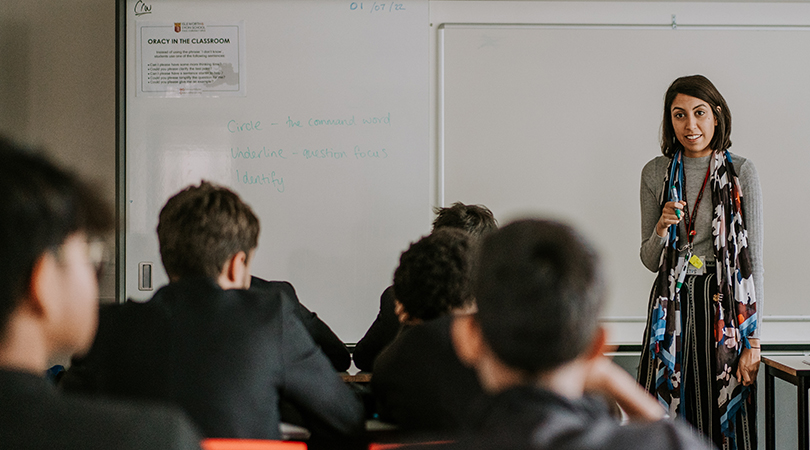 A female teacher stands at the front of a class, holding a pen.