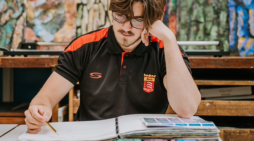 A Sixth Form student, dressed in a branded black polo, draws in his art book. 